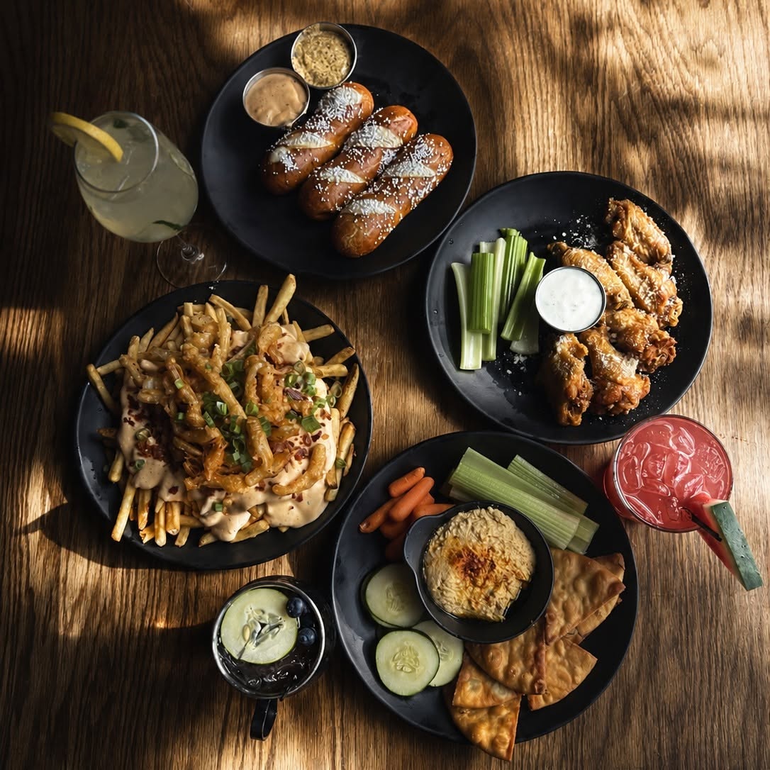 Overhead of a table spread in low sun: pretzels, loaded fries, wings, warm salmon dip and cocktails.